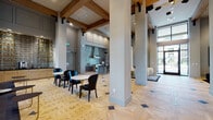 This elegantly designed single-floor amenity space makes an immediate impression with its dramatic, soaring ceilings accented by exposed wood beam detailing and bold structural columns. Entry is made through oversized navy steel-framed glass double doors, setting a refined tone that carries throughout the interior.

The herringbone-patterned hardwood flooring with decorative dark inlay accents anchors the space visually and flows seamlessly across all areas. Floor-to-ceiling steel-framed windows and glass partitions flood the interior with natural light while offering views of the surrounding landscape, including lush palm trees.

Multiple distinct seating zones offer varied atmospheres. A lounge area features a sculptural curved sofa, a marble coffee table, and a statement starburst chandelier centered above a fireplace with elegant wainscoting. A second casual lounge includes a fireplace, woven rattan accent chairs, and a textured area rug beneath exposed wood beam framing. A bar-style café nook showcases a dark granite counter with gold barstools, pendant lighting, and a curated shelving display.

A formal dining area is highlighted by a dark marble table, wishbone-style chairs, and two oversized gold drum pendant lights. A glass-walled office or conference room is visible beyond. A full kitchen with patterned tile backsplash, professional-grade appliances, and cabinetry completes the space, making it well-suited for both social gatherings and productive work.
