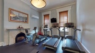 This fitness center offers a thoughtfully designed workout environment that seamlessly combines classic architectural character with comprehensive modern exercise equipment. The space is organized across two connected rooms, each finished with soft gray walls, crisp white crown molding, and white-trimmed doorways and windows that lend an elegant, refined aesthetic uncommon in typical gym settings.

The main cardio area is anchored by multiple treadmills and an elliptical trainer, complemented by a wall-mounted television for entertainment during workouts. Natural light filters in through wood-slatted blinds on tall windows, brightening the space alongside a distinctive drum pendant light fixture overhead. A wall-mounted split air conditioning unit ensures comfortable climate control year-round. A notable architectural centerpiece is the ornate marble fireplace, complete with a decorative mantel and framed artwork above, adding warmth and character to the room.

The adjoining strength training area provides a full rack of dumbbells in graduated weights, a flat weight bench, and a multi-function cable resistance machine. Rubber flooring throughout both rooms ensures safety and durability during exercise. A framed mirror on the wall of the weight room enhances the sense of space. The fitness center also features a paneled door providing access to an adjoining area, rounding out this versatile and well-appointed facility.