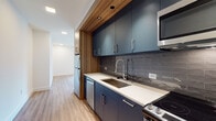 Located at 1150 Spring St NW in Atlanta's vibrant Midtown neighborhood, this single-floor unit delivers a clean, contemporary aesthetic throughout. Light-toned hardwood-style flooring runs continuously across the main living area, complementing the crisp white walls and recessed lighting overhead. A ceiling fan anchors the open living space, which receives generous natural light through large sliding glass doors that open to an outdoor view of the surrounding cityscape.

The galley-style kitchen is a standout feature, showcasing matte slate-blue cabinetry paired with warm wood ceiling accents, creating a striking contrast. Light quartz countertops, a glossy dark subway tile backsplash, and a deep stainless steel sink contribute to the refined look. Stainless steel appliances — including a refrigerator, dishwasher, range, and microwave — complete the functional layout.

A stylish barn-style door with frosted glass panels connects the living area to additional spaces, adding an industrial-modern touch. The bathroom features a matching dark-toned vanity with a white quartz countertop, undermount sink, and sleek wall-mounted lighting flanking a large mirror. Dark tile accents in the shower area echo the kitchen's design language, tying the space together with cohesive, modern detailing throughout.