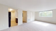 This single-floor unit is designed with a clean, neutral aesthetic throughout, featuring white walls, white trim, and light-toned carpeting that unifies the living spaces. The open-concept living area is generously sized, with natural light streaming in through a large window that frames views of surrounding greenery. A floor vent and dedicated closet provide practical storage and climate control within the main living space.

The kitchen is positioned adjacent to the living area and features dark espresso cabinetry, light-toned countertops, a stainless steel single-basin sink, and a dishwasher. The layout is efficient and functional, making good use of the available space.

Multiple bedrooms are carpeted and bathed in natural light from sizable windows, each offering views of mature trees. The rooms are well-proportioned, with clean walls and individual doors providing privacy. One bedroom includes a paneled white door leading to a closet.

The bathroom is finished in a bright, all-white palette, featuring a full tub-shower combination with white tile surround, chrome fixtures, a vanity with a white countertop, dark cabinetry below, and a wall-mounted mirror. The entry area is carpeted and opens directly into the main living space.