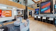 This stylish communal lounge is defined by its dramatic double-height ceilings and open, airy layout finished with light hardwood flooring throughout. Striking black glazed brick columns anchor the space vertically, creating visual separation between distinct activity zones while maintaining an open feel. The main lounge area features a tufted gold velvet sofa and contemporary grey accent chairs arranged around a sleek oval coffee table, with a large flat-screen TV mounted on a central display structure for entertainment. A bar-height counter with a dark granite surface and white bar stools occupies one corner, accompanied by a wall-mounted television and vibrant large-scale abstract artwork that adds bold color to the space. Built-in wood shelving units with glass accents display curated décor, contributing to the space's refined aesthetic. A shuffleboard table positioned near oversized windows allows natural light to flood that corner, offering views of the surrounding urban streetscape. A glass-walled private meeting or dining room features a chandelier and patterned wood accent wall, providing a more intimate setting within the broader space. A dramatic cityscape photographic mural crowns the upper portion of a wall, reinforcing the urban character of this well-appointed, multi-functional communal environment.