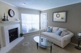 This single-floor space showcases a well-appointed open layout that connects the dining area and kitchen seamlessly. The dining space features carpeted flooring and is anchored by a round glass-top table paired with cream upholstered chairs, creating an intimate setting for meals. A two-tone wall treatment, combining warm brown and cream tones separated by white chair-rail molding, adds a classic decorative touch. Crown molding trims the ceiling throughout, contributing a refined architectural detail. A textured popcorn ceiling and a flush-mount ceiling light fixture provide functional overhead illumination. The adjacent kitchen area is partially visible, featuring a curved countertop overhang that forms a natural boundary between the two spaces, along with a range hood and standard cabinetry in warm wood tones. A wall-mounted HVAC vent ensures climate control throughout the space. The overall ambiance is warm and understated, with neutral tones creating a cohesive, inviting atmosphere suited to comfortable everyday living.