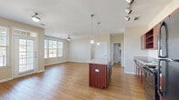 This single-floor, 1-bedroom, 1-bathroom residence offers a thoughtfully designed layout that maximizes both functionality and comfort. The open-concept kitchen and living area forms the heart of the home, featuring warm hardwood-style flooring, neutral beige walls, and large windows fitted with horizontal blinds that invite generous natural light. The kitchen is well-equipped with rich mahogany-toned cabinetry, granite-look countertops, black appliances including a range, microwave, dishwasher, and refrigerator, a double basin sink, and a central island with pendant lighting overhead. Track lighting adds additional illumination to the cooking space. A door with transom window provides access to an outdoor area from the dining space. The spacious bedroom is carpeted for warmth and comfort, features a ceiling fan, and benefits from natural light through large windows. It connects directly to the full bathroom and an adjacent closet. The bathroom includes a tub-shower combination, a mahogany vanity with granite-look countertop, and hardwood-style flooring. Ceiling fans are present throughout the main living areas, contributing to a comfortable and practical living environment.