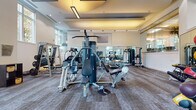 This fitness center is a comprehensively outfitted exercise facility designed to support a wide range of workout routines. The space is organized into distinct functional zones, each dedicated to a specific category of training. The cardio section features a row of treadmills, elliptical trainers, and upright stationary bikes, all positioned along mirrored walls that visually expand the room. A wall-mounted television provides entertainment during workouts.

The strength training area includes a multi-station cable machine offering numerous exercise configurations, along with a power rack and weight bench for barbell training. A dedicated free weights station houses a full rack of dumbbells in graduated sizes, accompanied by stability and medicine balls for functional fitness exercises.

Natural light floods the space through large, floor-to-ceiling windows that overlook a tree-lined streetscape, creating an open and energizing atmosphere. The ceiling features a recessed tray design with warm pendant and recessed lighting, complementing the abundant daylight. Full-length mirrors line multiple walls, enhancing the sense of space throughout.

The flooring consists of durable, textured carpet tiles suited for both cardio and strength activities. The overall design is clean and modern, with white walls and a well-organized layout that ensures efficient use of the available space.