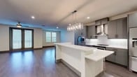 This two-story residence is designed with a clean, contemporary aesthetic, combining functionality with modern style across both levels. The main living area is anchored by an open-concept layout that seamlessly connects the kitchen, dining, and living spaces. The kitchen is a standout feature, equipped with stainless steel appliances, a stainless range hood, gray cabinetry with matte black hardware, and a large white quartz island topped with a striking multi-light pendant chandelier. A built-in wine cooler and additional gray cabinetry line the entryway corridor, adding practical storage with a polished look. Hardwood-style flooring extends throughout the main living areas, complementing the neutral wall tones. French doors with slatted blinds open to a balcony, allowing natural light to flow into the living space, which is also served by a ceiling fan. The upper level features an open landing with built-in shelving and a ceiling fan. The primary bathroom offers a double-sink vanity with gray cabinetry, a walk-in tiled shower, and light tile flooring. A generously sized walk-in closet provides ample built-in shelving and hanging rods. Additional rooms feature carpet flooring. The oversized attached garage accommodates multiple vehicles with ease.