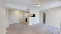 This single-floor residence offers a clean, open-concept layout designed for comfortable everyday living. The living area is finished with wall-to-wall neutral carpeting, creating a warm and cohesive atmosphere throughout the main space. A defining feature of the layout is the kitchen peninsula, which serves as a natural divider between the kitchen and living area while maintaining an open, connected feel. The kitchen is equipped with white cabinetry, a built-in sink, and dark appliances, including what appears to be a refrigerator and dishwasher. Light-toned flooring in the kitchen contrasts neatly with the carpeted living space, adding visual definition to each zone. The walls are finished in a soft, neutral white with subtle warm undertones, contributing to a bright and airy ambiance. Overhead lighting is provided by a flush-mount ceiling fixture, and HVAC ventilation is integrated into the ceiling for climate comfort. A doorway visible from the living area leads to additional rooms, suggesting a practical and well-organized floor plan. The overall aesthetic is understated and functional, with clean lines and a neutral palette that offers a versatile backdrop for a variety of interior styles.