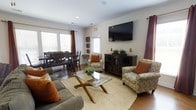 Located at 100 Durham Park Way in Pooler, Georgia, this single-floor community space offers a warm and inviting environment designed for comfort and social gathering. The layout seamlessly blends a lounge area with a dining space, creating a cohesive and functional setting.

The lounge area is elegantly appointed with patterned accent chairs featuring burnt orange throw pillows, a plush gray sofa, and a glass-top coffee table set atop a textured woven area rug. A wall-mounted flat-screen television serves as a focal point, complemented by a dark wood media console with glass-door cabinetry below. Decorative greenery and table lamps add a finishing touch to the space.

Adjacent to the lounge, a dining area accommodates a dark wood table with matching X-back chairs, ideal for group gatherings. Large windows dressed with rich burgundy drape panels allow natural light to fill the room, enhancing the warm, neutral color palette throughout.

Crown molding and recessed lighting contribute to the polished, well-finished feel of the interior. The hardwood-style flooring ties the spaces together, providing both durability and aesthetic appeal. Overall, this space strikes a balance between casual comfort and refined design, making it well-suited for residents and visitors alike.