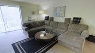 This single-floor space features a comfortable and well-appointed living area designed for relaxed everyday living. The room is anchored by a large, wrap-around sectional sofa in a warm taupe fabric, offering generous seating and a casual, inviting feel. A round wooden coffee table with a glass top sits at the center, positioned atop a bold black-and-white striped area rug that adds a graphic contrast to the neutral carpeted flooring. The walls are painted in a soft, light green tone, contributing to the room's calm and airy ambiance. Decorative artwork, including a botanical floral canvas, adds a touch of color and personality to the walls. A tall arc floor lamp provides warm ambient lighting, complementing the natural light that filters in through what appears to be a sliding glass door or large window along one wall. A wall-mounted air conditioning unit suggests climate control convenience. Overall, the living area balances comfort and functionality, making it a practical and welcoming space for everyday use.