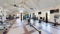 Located in Lady Lake, Florida, this single-floor fitness center offers a generously sized, thoughtfully designed workout facility that caters to a wide range of exercise needs. The space is defined by its striking vaulted ceiling with exposed white beams, which rises dramatically overhead to create an open, airy atmosphere. Multiple large-blade ceiling fans are suspended from the peak, ensuring comfortable airflow throughout. Recessed lighting complements the abundant natural light that streams in through large windows lining the perimeter walls, providing bright, welcoming conditions at any time of day.

The main workout floor features a distinctive checkered tile pattern in contrasting light and dark tones, lending the space a clean, modern aesthetic. The primary area is outfitted with a comprehensive selection of strength training equipment, including cable machines, selectorized weight stations, a power rack, benches, and a full dumbbell rack displayed in front of a wall-length mirror. The cardio zone houses treadmills, ellipticals, stair climbers, and rowing machines. A separate functional training room accommodates battle ropes, stability balls, medicine balls, and resistance accessories, offering a dedicated space for dynamic and functional workouts. The overall ambiance is bright, professional, and motivating.
