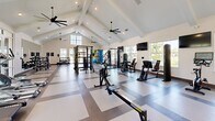 Located in Lady Lake, Florida, this single-floor fitness center offers a generously sized, thoughtfully designed workout facility that caters to a wide range of exercise needs. The space is defined by its striking vaulted ceiling with exposed white beams, which rises dramatically overhead to create an open, airy atmosphere. Multiple large-blade ceiling fans are suspended from the peak, ensuring comfortable airflow throughout. Recessed lighting complements the abundant natural light that streams in through large windows lining the perimeter walls, providing bright, welcoming conditions at any time of day.

The main workout floor features a distinctive checkered tile pattern in contrasting light and dark tones, lending the space a clean, modern aesthetic. The primary area is outfitted with a comprehensive selection of strength training equipment, including cable machines, selectorized weight stations, a power rack, benches, and a full dumbbell rack displayed in front of a wall-length mirror. The cardio zone houses treadmills, ellipticals, stair climbers, and rowing machines. A separate functional training room accommodates battle ropes, stability balls, medicine balls, and resistance accessories, offering a dedicated space for dynamic and functional workouts. The overall ambiance is bright, professional, and motivating.