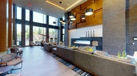 This modern lobby lounge is defined by its dramatic double-height ceilings and expansive floor-to-ceiling windows that flood the space with natural light and offer views of the surrounding greenery. The architectural design blends warm wood wall paneling with cool stone and dark brick accents, creating a striking visual contrast that anchors the room's contemporary aesthetic. A wide, linear gas fireplace serves as the focal point of one wall, framed by a sculptural stone surround and flanked by a built-in log storage niche, adding both warmth and visual interest. A curated arrangement of cylindrical pendant lights hang at varying heights from the ceiling, providing layered illumination and reinforcing the space's vertical grandeur. Generous seating is provided through plush, low-profile sofas and lounge chairs in neutral tones, accented with bold yellow cushions that add a vibrant pop of color. A multicolored striped area rug defines the central seating zone and ties the palette together. Large-format light-toned floor tiles extend throughout, contributing to the clean, open feel of the space. Additional seating areas near the windows offer quieter, light-filled spots ideal for relaxation or informal gatherings, completing this well-appointed and inviting lounge environment.