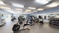 This spacious, single-floor fitness center is thoughtfully designed to accommodate a comprehensive workout experience. The facility is finished with durable rubber flooring throughout, blue-painted walls, and a textured ceiling lined with bright recessed fluorescent lighting that ensures the space is well-lit at all times. Large windows along multiple walls invite natural light and provide pleasant views of the surrounding greenery, creating an open and energizing atmosphere.

The cardio zone features treadmills, stationary bikes, and elliptical machines, with wall-mounted televisions nearby for entertainment during workouts. A water cooler station is conveniently positioned near the entrance area, which also provides glass door access to an adjacent outdoor area.

The strength training section is extensively equipped, offering a Cybex power rack with a adjustable bench, a multi-station cable machine, barbells with weight plates, and a full rack of dumbbells. Additional selectorized machines are arranged throughout the space, catering to a variety of muscle groups. Full-length mirrors line one wall, allowing users to monitor their form during exercises. Stability balls and other accessories round out the offerings, making this fitness center a versatile and fully functional facility suitable for users of all fitness levels.