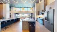 Located at 12888 Crenshaw Blvd in Gardena, CA, this single-floor communal amenity space offers a thoughtfully designed environment for socializing and recreation. The interior is anchored by a striking kitchen featuring a large terrazzo-clad island with a stone countertop, wire bar stools, wood-tone cabinetry, white subway tile backsplash, professional-grade stainless appliances, and a sleek range hood. Soaring double-height ceilings with bold geometric LED lighting and globe pendant lights add dramatic architectural flair throughout. An expansive lounge area includes a full-size billiards table set atop terrazzo flooring, a mounted flat-screen TV, and an orange upholstered banquette. Tucked along one side are intimate private study or work booths lined in teal textured fabric, each equipped with task lighting and small desks. Floor-to-ceiling glass bi-fold doors seamlessly connect the interior to a spacious outdoor patio, where multiple shaded dining tables and woven chairs are arranged for al fresco gatherings. Lush planted borders and open sky views complete the outdoor setting. The hallway and entrance transition zones are clean and modern, ensuring a polished flow throughout this well-appointed communal retreat.