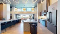 Located at 12888 Crenshaw Blvd in Gardena, CA, this single-floor communal amenity space offers a thoughtfully designed environment for socializing and recreation. The interior is anchored by a striking kitchen featuring a large terrazzo-clad island with a stone countertop, wire bar stools, wood-tone cabinetry, white subway tile backsplash, professional-grade stainless appliances, and a sleek range hood. Soaring double-height ceilings with bold geometric LED lighting and globe pendant lights add dramatic architectural flair throughout. An expansive lounge area includes a full-size billiards table set atop terrazzo flooring, a mounted flat-screen TV, and an orange upholstered banquette. Tucked along one side are intimate private study or work booths lined in teal textured fabric, each equipped with task lighting and small desks. Floor-to-ceiling glass bi-fold doors seamlessly connect the interior to a spacious outdoor patio, where multiple shaded dining tables and woven chairs are arranged for al fresco gatherings. Lush planted borders and open sky views complete the outdoor setting. The hallway and entrance transition zones are clean and modern, ensuring a polished flow throughout this well-appointed communal retreat.