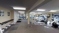 This single-floor fitness center offers a practical and functional space designed to support a well-rounded exercise routine. The open layout provides generous room to move between equipment, with a carpet and rubber mat flooring combination that balances comfort and safety underfoot.

The facility is outfitted with a solid selection of cardio machines, including multiple treadmills positioned near large windows that invite natural light and offer outdoor views — ideal for keeping workouts engaging. Strength and resistance training equipment occupies the central area of the room, including cable and weight machines suitable for a range of exercises.

Overhead, flush-mounted fluorescent lighting ensures the space is consistently and evenly illuminated throughout, complementing the natural light streaming in from the surrounding windows. A central support column divides the floor plan subtly without interrupting the overall sense of openness.

The neutral color palette of cream walls and a gray ceiling contributes to a clean, no-frills aesthetic that keeps the focus on functionality. A glass-panel entry door at the far end adds to the brightness of the space. Whether used for cardio, strength training, or stretching, this fitness center provides a well-lit, organized, and accessible environment for a comprehensive workout experience.