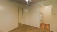 This single-floor space is characterized by neutral, light-colored walls that create a clean and straightforward aesthetic throughout. The main living area features wall-to-wall carpet in a warm beige tone, providing a comfortable underfoot surface. A ceiling fan is centrally mounted, offering both lighting and air circulation, while an overhead HVAC vent ensures climate control throughout the space.

A mirrored sliding closet door is positioned along one wall, adding functional storage while reflecting light to enhance the sense of openness in the room. The adjacent kitchen area transitions to hardwood-style flooring, providing a practical and easy-to-maintain surface. Visible kitchen appliances include a white refrigerator and what appears to be a white range, indicating a fully equipped cooking space.

The layout appears open and straightforward, with clear sightlines between the main living area and the kitchen. Neutral wall tones throughout the space offer flexibility for personalization with furnishings and décor. The overall ambiance is modest and functional, suited for everyday living.