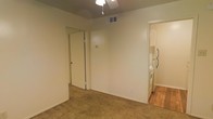 This single-floor space is characterized by neutral, light-colored walls that create a clean and straightforward aesthetic throughout. The main living area features wall-to-wall carpet in a warm beige tone, providing a comfortable underfoot surface. A ceiling fan is centrally mounted, offering both lighting and air circulation, while an overhead HVAC vent ensures climate control throughout the space.

A mirrored sliding closet door is positioned along one wall, adding functional storage while reflecting light to enhance the sense of openness in the room. The adjacent kitchen area transitions to hardwood-style flooring, providing a practical and easy-to-maintain surface. Visible kitchen appliances include a white refrigerator and what appears to be a white range, indicating a fully equipped cooking space.

The layout appears open and straightforward, with clear sightlines between the main living area and the kitchen. Neutral wall tones throughout the space offer flexibility for personalization with furnishings and décor. The overall ambiance is modest and functional, suited for everyday living.
