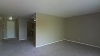 This single-floor residence offers a straightforward and functional layout designed for comfortable everyday living. The living area is finished with wall-to-wall carpeting in a neutral tone, creating a warm and quiet underfoot experience. Smooth, light-colored walls with white baseboards give the space a clean and understated aesthetic throughout.

A partial wall divides the living area from the kitchen, which is visible through an open passageway. The kitchen features cabinetry in a warm wood finish, standard appliances, and a ceiling fan that provides ventilation and air circulation. The transition between the two areas is seamless, supporting an open and connected feel between the main living and cooking spaces.

The textured ceiling adds a subtle decorative element consistent throughout the home. The neutral color palette of beige and gray tones creates a cohesive and versatile backdrop, making the space easy to personalize. Overall, the residence presents a practical and livable environment with a modest, no-frills design that prioritizes function and comfort.
