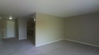 This single-floor residence offers a straightforward and functional layout designed for comfortable everyday living. The living area is finished with wall-to-wall carpeting in a neutral tone, creating a warm and quiet underfoot experience. Smooth, light-colored walls with white baseboards give the space a clean and understated aesthetic throughout.

A partial wall divides the living area from the kitchen, which is visible through an open passageway. The kitchen features cabinetry in a warm wood finish, standard appliances, and a ceiling fan that provides ventilation and air circulation. The transition between the two areas is seamless, supporting an open and connected feel between the main living and cooking spaces.

The textured ceiling adds a subtle decorative element consistent throughout the home. The neutral color palette of beige and gray tones creates a cohesive and versatile backdrop, making the space easy to personalize. Overall, the residence presents a practical and livable environment with a modest, no-frills design that prioritizes function and comfort.