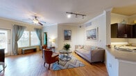 This single-floor residence showcases a thoughtfully designed layout with a coastal aesthetic that flows seamlessly from room to room. The living area serves as the heart of the home, featuring warm hardwood flooring, crown molding, and a ceiling fan overhead. Track lighting complements the natural light filtering through large windows dressed in teal curtains. A glass-top coffee table, upholstered sofa, and accent chairs rest atop a chevron-patterned area rug, while nautical-themed artwork adorns the walls. Adjacent to the living space, a cozy dining nook accommodates a round glass-top table with wicker seating, framed by additional coastal artwork. The kitchen is well-equipped with dark espresso cabinetry, light countertops, a white subway tile backsplash, and a full suite of black appliances, including a range, over-the-range microwave, and refrigerator. Two carpeted bedrooms offer a serene retreat, each featuring ceiling fans, natural light, closet access, and beach-inspired wall art. The primary bathroom presents a dual-sink vanity with a stone-toned countertop, dark cabinetry, a large mirror, and a glass-enclosed walk-in shower with decorative tile accents. A second bathroom mirrors the refined finishes with a single-sink vanity and tiled flooring.