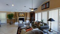 Located on Loudon Court in Fayetteville, North Carolina, this single-floor home offers a well-designed layout with a warm and inviting atmosphere. The living room serves as the centerpiece of the home, showcasing soaring vaulted ceilings that lend a sense of openness and grandeur. A classic fireplace with a dark surround anchors one wall, creating a natural focal point and adding a cozy, welcoming feel to the space. Large windows dressed with vertical blinds line the exterior wall, allowing generous amounts of natural light to pour in and brighten the warm, neutral-toned walls. A ceiling fan with a light fixture hangs centrally from the vaulted ceiling, providing both functional airflow and a finishing architectural touch. The living area is generously proportioned, comfortably accommodating multiple seating arrangements including plush sofas and accent chairs centered around a glass-top coffee table atop a decorative area rug. A dark wood bookcase adds both storage and character to the room, complementing the overall earthy, traditional aesthetic. Recessed and track lighting provide additional illumination throughout. The overall ambiance of the home is warm, classic, and comfortable, making it a well-suited residence in the Fayetteville area.