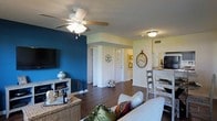 This single-floor residence offers a thoughtfully designed open-concept layout that connects the living, dining, and kitchen areas in a cohesive and functional arrangement. The living room makes a striking first impression with a bold teal accent wall that serves as a vibrant backdrop for the wall-mounted flat-screen television and a white media console. Rich, dark hardwood flooring runs throughout, adding warmth and continuity to the space. A ceiling fan with an integrated light fixture provides both comfort and illumination, complementing the additional flush-mount ceiling lights that keep the space bright and welcoming. Decorative wall accents, including a large ornamental clock, lend personality and character to the interior. The living area flows naturally into a dining space furnished with a light-toned table and wooden chairs, positioned conveniently adjacent to the kitchen. The kitchen is visible in the background and features dark appliances. A hallway leads to additional rooms beyond, with multiple interior doors hinting at well-defined private spaces. The textured ceiling and neutral wall tones throughout provide a balanced contrast to the bold design choices, resulting in a space that is both stylish and inviting.