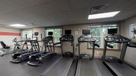 This fitness center is organized across a single floor and offers a comprehensive range of exercise options within a clean, functional layout. The cardio area is lined with multiple treadmills and elliptical trainers, each equipped with individual monitor screens, and is bathed in natural light from large windows overlooking lush greenery — creating a refreshing atmosphere for workouts. Rubber flooring throughout the fitness zones provides both comfort and durability underfoot.

The strength training area is distinctly styled with bold red accent stripes along neutral gray walls, accented by motivational typography decals. It houses a multi-function cable machine, a power rack with an adjustable bench, a full dumbbell rack, and open floor space ideal for functional training. Wall-mounted mirrors enhance the sense of space and allow users to monitor their form. Exercise balls and medicine balls are also available for dynamic training.

A separate, dedicated room serves as a bike storage and maintenance station, complete with a bike repair stand, floor pump, and secure bike racks — a thoughtful amenity for cycling enthusiasts. Recessed and panel lighting ensures consistent illumination throughout all areas. The facility's well-organized design and varied equipment make it a versatile space suited for a wide range of fitness routines and training styles.