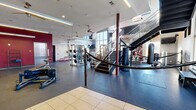 Located on West Tennessee Street in Tallahassee, FL, this two-floor fitness facility offers a generous and well-equipped workout environment spread across a large main floor and a secondary upper level. The main floor commands an impressive presence with soaring ceilings, exposed structural columns, and abundant natural light streaming through expansive floor-to-ceiling windows. The rubber flooring throughout the workout areas provides both durability and comfort underfoot, transitioning to tile in the entry and corridor zones. A bold red and white color scheme energizes the space, reinforced by accent walls and branded signage. The main floor houses a comprehensive free weight section featuring a full rack of dumbbells alongside wall-length mirrors, multiple cable and resistance machines, weight benches, and functional training equipment including battle ropes and a heavy punching bag within a dedicated boxing corner. A row of treadmills and stationary bikes lines one section, catering to cardio-focused workouts. Ceiling fans are mounted throughout to aid ventilation. A metal staircase with open railings leads to the upper level, which provides additional workout space overlooking the main floor. Restrooms and support spaces are accessible via a tiled corridor. The facility's open layout and high-visibility design make it a functional and motivating training environment.