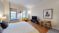 Located in Boston's vibrant Chinatown neighborhood, this single-floor studio at 27 Beach St offers a thoughtfully designed open layout that efficiently combines living, sleeping, and dining functions. The main living area features warm hardwood flooring, crown molding, and crisp white walls that create a bright and inviting atmosphere. Natural light filters in through multiple windows, enhancing the sense of openness within the space.

The sleeping area is anchored by a generously sized bed positioned against a textured accent wall, complemented by warm ambient lighting and framed artwork. A flat-screen TV, a mustard yellow accent chair, and a compact dining table with chairs complete the living portion of the room, providing comfort and functionality in a cohesive arrangement.

The kitchenette is neatly tucked into a dedicated alcove and features rich dark cabinetry, granite countertops, a subway tile backsplash, a stainless steel sink, and a full range — offering all the essentials for in-unit cooking. The bathroom includes a granite-topped vanity, large mirror, and a tiled shower enclosure with neutral tones, maintaining a clean and functional aesthetic throughout.