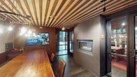 This conference room offers a refined yet inviting atmosphere, blending functional design with striking architectural details. The ceiling is a standout feature, showcasing a slatted wood pattern that adds warmth and visual texture overhead, complemented by track lighting that allows for flexible illumination throughout the space.

A built-in gas fireplace set within a floor-to-ceiling tiled surround serves as a dramatic focal point along one wall, lending the room an ambiance that feels both sophisticated and comfortable. The dark, elongated tile cladding surrounding the fireplace provides a bold contrast to the warmer wood tones found elsewhere in the room.

A long solid wood conference table anchors the space, accompanied by wooden chairs that reinforce the room's cohesive, natural aesthetic. A large wall-mounted display screen is positioned for easy viewing during presentations or collaborative sessions.

Glass doors at the far end of the room introduce natural light and provide a visual connection to an adjacent area, preventing the space from feeling enclosed. Dark hardwood flooring ties all the elements together, contributing to an overall atmosphere that is professional, warm, and uniquely designed.