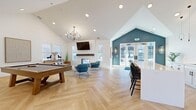 This single-floor communal space offers a well-appointed, open-concept layout designed for socializing and relaxation. The interior is immediately distinguished by soaring vaulted ceilings that amplify the sense of volume throughout, complemented by recessed lighting and a striking multi-arm chandelier that anchors the main lounge area.

The entry foyer features glass-paneled French doors set against a reclaimed wood accent wall, flanked by pendant wall sconces and framed by herringbone-patterned light hardwood flooring that extends seamlessly across the entire space. A welcoming seating area near the entrance transitions into the main gathering zone.

The lounge area centers on a shiplap fireplace with a rustic wood beam mantel and wall-mounted television, surrounded by contemporary seating including a leather sofa and accent chairs. A billiards table occupies a prominent central position, encouraging recreational use. Curated artwork lines one wall, adding a gallery-like quality.

A dedicated kitchenette features white cabinetry, a marble-topped island with bar seating, a beverage refrigerator, and open floating shelves. A teal accent wall near the secondary entrance adds a bold architectural focal point. Natural light flows generously through multiple windows and glass doors, enhancing the bright, modern ambiance throughout.