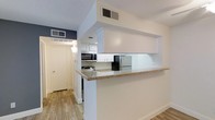 This single-story space showcases a thoughtfully designed kitchen that serves as a central feature of the layout. The kitchen is framed by crisp white cabinetry that extends to the upper walls, providing ample storage while maintaining a clean, bright aesthetic. A peninsula counter finished in granite with warm brown and beige tones creates a natural divide between the kitchen and the adjacent living area, offering both additional workspace and a casual seating option.

The kitchen is equipped with stainless steel appliances, including a refrigerator, microwave, and range, complementing the modern cabinetry. A built-in overhead range hood adds a structured architectural element above the cooking area. The textured ceiling adds subtle character throughout the space.

Wood-look flooring runs continuously through the kitchen and into the hallway, lending warmth and visual continuity to the interior. The hallway connects to an entry door and additional rooms, with wall-mounted lighting providing a welcoming ambiance. Multiple overhead HVAC vents are positioned throughout, ensuring consistent climate control. The overall design balances functionality with a clean, contemporary aesthetic.