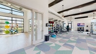 This contemporary fitness center is thoughtfully designed to support a wide range of exercise routines across multiple distinct zones. The main workout area features a striking geometric-patterned floor in soft blues and greens, soaring ceilings with bold dark exposed beams, and pendant lighting that adds a modern industrial touch. Ceiling fans complement the open, airy atmosphere throughout.

The strength training zone is comprehensively outfitted with a cable machine system, a full rack of kettlebells, medicine balls, barbells, and weight benches, all arranged alongside large wall-mounted mirrors. An adjacent cardio section lines multiple treadmills and ellipticals along a wall of oversized windows, offering views of lush palm trees and landscaping that bring the outdoors in.

A separate, dedicated studio space features light hardwood flooring, soaring double-height ceilings, floor-to-ceiling black-framed windows that flood the room with natural light, and a full mirrored wall paired with a large display screen—ideal for group fitness classes, yoga, or personal training sessions. Resistance bands, stability balls, and additional accessories are neatly stored within the studio.

Throughout the facility, the bright white walls, generous glazing, and clean architectural lines create an energizing and welcoming environment well-suited for a comprehensive fitness experience.