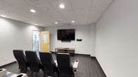 This single-floor screening room is purpose-built for a premium media viewing experience, combining functional design with a polished, modern aesthetic. The space is anchored by a large wall-mounted flat-screen display paired with a soundbar and accompanying media console, creating a focused entertainment setup along the primary wall.

Seating is arranged in theater-style rows, featuring distinctive blue and white two-tone reclining chairs equipped with built-in cup holders and wide armrests. The chairs are designed for comfort during extended viewing sessions and are arranged to provide clear sightlines to the display from every position in the room. A structural column near the center-rear of the space subtly divides the back seating area without disrupting the overall layout.

The room's interior is finished with neutral light-gray walls and dark tile flooring laid in a linear pattern, lending a clean and contemporary feel. A drop tile ceiling with recessed can lighting provides even, controlled illumination throughout. A yellow-accented entry door leads to an adjacent area visible through a glass panel, suggesting the room is part of a larger facility. The overall ambiance is modern, functional, and well-suited for group screenings, presentations, or communal media viewing.