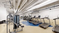 This expansive fitness center offers a comprehensive workout facility across a single open floor, thoughtfully organized into distinct training zones. The space is immediately recognizable by its striking two-tone rubberized flooring — bold blue zones contrast with neutral beige areas — providing both visual definition and practical cushioning underfoot.

The cardio zone is well-stocked with a row of treadmills and elliptical machines, each equipped with individual digital displays, alongside spin bikes for cycling workouts. Strength training is extensively covered, with a dedicated free weights area featuring a full rack of dumbbells, kettlebells, medicine balls, and a barbell bench press station with weight plates. A multi-functional cable machine anchors a separate functional training zone, complemented by TRX suspension systems, punching bags, and plyo boxes.

Additional selectorized strength machines are distributed throughout the space, offering targeted muscle group training. Wall-mounted mirrors in the free weights area aid in form and technique. The ceiling features exposed ductwork, piping, and overhead fluorescent lighting, giving the facility an industrial yet clean and bright aesthetic. The white walls and bright lighting ensure the space feels open and well-illuminated, creating an energizing environment for a full range of fitness activities.