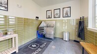 Located at 411 Sage Rd N in White House, Tennessee, this purpose-built pet grooming room — branded as the "Dog Spa" — is a thoughtfully designed, single-room amenity that combines functionality with a clean, polished aesthetic. The space is finished with distinctive olive-green glazed subway tile that wraps the lower half of all four walls, creating a durable, easy-to-clean surface while adding a warm, stylish character to the room. Dark slate-style floor tiles provide a practical and attractive foundation underfoot. Natural light floods the interior through multiple large windows, including a set of three tall windows along one wall that offer views of the surrounding residential community, and an additional window flanking a glass-paneled exterior door for easy access. The grooming area is well-equipped, featuring a stainless steel grooming tub with an integrated faucet and tool storage, wall-mounted dispensers for grooming products, a freestanding pet dryer on a rolling stand, and a wooden bench for pet owners. A light wood utility table holds treat jars and supplies, adding a welcoming touch. Three framed dog-themed art prints adorn the upper wall, reinforcing the space's dedicated canine focus. A floor mat emblazoned with "Dog Spa" anchors the room's identity, making this a fully outfitted and inviting grooming destination.