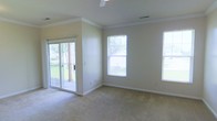 This single-floor space offers a clean, well-lit interior defined by classic architectural details and a functional layout. The main living area is finished with wall-to-wall carpeting in a neutral tone, complementing the soft beige walls and white crown molding that frames the ceiling throughout. A ceiling fan provides both comfort and airflow overhead.

Natural light fills the room through multiple windows, each fitted with white trim that ties cohesively into the overall color scheme. The windows offer views of the surrounding outdoor greenery, creating a pleasant connection to the exterior environment. A sliding glass door provides direct access to an outdoor deck or patio, further enhancing the sense of openness and allowing abundant natural light to penetrate the interior.

The space features white baseboards and recessed HVAC vents integrated into the ceiling, reflecting a practical and tidy design approach. The neutral palette of beige walls and cream-toned trim creates a versatile backdrop suitable for a variety of furnishing styles. Overall, the space presents a bright, airy ambiance with traditional detailing and comfortable proportions throughout.