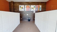 This single-floor pet grooming and wash station is thoughtfully designed to accommodate pet owners with style and functionality. The space is divided into distinct zones, beginning with a bright, glass-walled entry featuring floor-to-ceiling windows and black-framed glass doors that flood the interior with natural light and offer views of a landscaped outdoor courtyard and surrounding community buildings. A bold orange mosaic tile accent wall anchors the entry area, where a mounted pet dryer and a bank of sleek white high-gloss storage lockers provide convenient access to grooming supplies and personal belongings. The main wash bay is a standout feature, finished with wood-look wall tile that lends a warm, modern aesthetic. Flanking walls are lined with white cabinetized panels, and the floor is covered in small-format neutral mosaic tile with a centrally located floor drain for easy cleanup. A series of four colorful, pop-art-style dog portraits adorns the upper wall, adding a playful and welcoming character to the space. Recessed lighting and a ventilation system overhead ensure a comfortable environment. The overall design balances practicality with personality, making this a well-appointed amenity for pet owners.