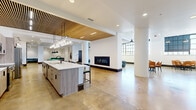 Located at 712/2 Pearl St in Sioux City, Iowa, this expansive club room offers a generously sized, open-plan gathering space designed with a contemporary aesthetic and a variety of amenity zones. Polished concrete floors flow throughout, providing a sleek, industrial-modern foundation that ties the entire space together.

The kitchen area anchors one section of the room, featuring a large island with a white quartz countertop, wood-toned cabinetry, and an elegant hanging wine glass rack overhead. A striking wood slat ceiling detail with integrated lighting frames this zone, adding warmth and visual interest. A linear gas fireplace is set into a crisp white wall, creating a natural focal point for the adjacent lounge area, which is furnished with curved teal accent chairs.

Large steel-framed industrial windows along one wall flood the space with natural light and lend a loft-inspired character. The seating areas are thoughtfully arranged with a mix of round dining tables paired with warm orange chairs, intimate booth seating with teal upholstery, and additional lounge clusters. A dedicated billiards area with a full-size pool table and pendant lighting rounds out the recreational offerings. Tall white columns punctuate the open floor plan, while curated artwork and carefully chosen furnishings complete the polished, community-focused atmosphere.