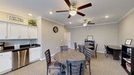 Located at 890 Quail Creek Blvd in Stow, Ohio, this single-floor community space is thoughtfully designed to accommodate social gatherings and everyday convenience. The central dining area serves as the heart of the space, featuring warm neutral carpeting, crown molding, and dual ceiling fans with integrated lighting that provide both comfort and style. Multiple round dining tables with ladder-back chairs offer flexible seating arrangements, while a built-in kitchenette with white cabinetry and a dark countertop with sink provides practical refreshment support. A dedicated nook within the dining area houses a bookshelf stocked with books and games, along with a small desk, creating a quiet corner for leisure activities. Natural light flows generously through large windows fitted with blinds, and elegant French doors with transom windows lead to an adjacent area, enhancing the open and connected feel of the space. The hallway transitions seamlessly between areas, finished with durable wood-look vinyl plank flooring that extends into both bathrooms. Each bathroom is fully ADA-compliant, featuring large-format neutral tile walls, wood-look vinyl flooring, stainless steel grab bars, and commercial-grade fixtures, ensuring accessibility and ease of use for all visitors. The overall ambiance is clean, welcoming, and functional.