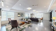 Located at 205 W Touhy Ave in Park Ridge, Illinois, this thoughtfully designed single-floor community space offers a sophisticated and welcoming environment. The main lounge area is expansive, featuring rich dark hardwood flooring, recessed lighting, and flush drum pendant fixtures that cast a warm, even glow throughout. Comfortable seating arrangements — including plush sofas, accent chairs, and a tufted leather ottoman — are complemented by a decorative console table, a striking circular mirror, and built-in shelving, creating an inviting atmosphere for socializing or relaxing. Large windows flood the space with abundant natural light and offer views of the surrounding greenery. Connecting hallways continue the clean, modern aesthetic with light hardwood floors, built-in bench seating, and generous window placements. The dining room is a standout feature, distinguished by a tray ceiling, textured wall coverings, and a glamorous crystal chandelier. Floor-to-ceiling cabinetry in a warm wood and slate blue finish provides both display and storage, while a large dining table surrounded by upholstered chairs makes it ideal for private gatherings. Elegant black-framed glass partition doors connect the dining area to the lounge, adding an architectural flourish that enhances the overall sense of refined, contemporary design.