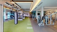 Located along the waterfront at 150 W Ocean Blvd in Long Beach, California, this state-of-the-art fitness center offers a comprehensive workout environment designed for a wide range of training styles. The interior is thoughtfully designed with teal accent walls, coffered ceilings with warm ambient yellow lighting, and large floor-to-ceiling sliding glass doors that flood the space with natural light and provide seamless access to an outdoor terrace. The main training floor features a turf track lined with yard markers, ideal for functional and athletic training. A diverse array of equipment is on hand, including strength machines, free weights with a full dumbbell rack, a RAX functional training rig, a StairMaster, and a heavy punching bag. A striking large-scale black-and-white mountain mural serves as a dramatic focal point on one wall, adding an artistic dimension to the space. Accent shelving in bold yellow tones displays medicine balls and accessories. The outdoor area extends the fitness experience with cycling bikes positioned under a pergola-style canopy, lounge seating, and poolside views framed by swaying palm trees, creating a resort-like atmosphere. Together, the indoor and outdoor zones form a comprehensive, modern fitness facility.