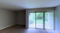 This single-floor space presents a clean, functional layout with a focus on connecting the interior to the outdoors. The main living area is finished with wall-to-wall carpeting in a neutral tone, complementing the gray-painted walls and white ceiling to create a calm, understated atmosphere. A prominent sliding glass door serves as the room's focal point, fitted with vertical blinds that allow for adjustable light control. The door opens directly onto a patio area, beyond which mature trees and dense green foliage provide a naturally private and serene outdoor backdrop. Natural light filters generously into the room through this large glazed opening, brightening the interior throughout the day. The overall design is straightforward and practical, with smooth walls and standard ceiling height contributing to a tidy, open feel. The neutral color palette throughout offers a versatile canvas well-suited to a variety of furnishing styles. The seamless connection between the indoor living space and the verdant outdoor surroundings is a notable characteristic of this unit, enhancing the sense of openness and tranquility within the space.
