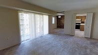 This single-floor residence offers an open and airy layout filled with natural light. The living area is spacious, anchored by large floor-to-ceiling sliding glass doors dressed with vertical blinds that open to a balcony, allowing ample daylight to flood the carpeted interior. The neutral-toned walls and light carpet throughout create a clean, cohesive aesthetic.

A standout architectural feature is the exposed brick accent wall that separates the living space from the adjoining dining or secondary area. This warm, textural element adds character and contrast to the otherwise understated interior. A pendant light fixture hangs above this transitional space, providing both functional and ambient lighting.

The layout flows naturally between the main living area and the connected secondary space, suggesting versatility in how the rooms can be used. A louvered door is visible toward the rear, likely leading to a utility or storage area, and additional doorways hint at further rooms beyond the main living zone. Overall, the space offers a balanced combination of functional design and inviting architectural detail.
