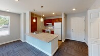 This single-floor unit in Kokomo, Indiana offers a thoughtfully designed, open layout with a neutral color palette of soft gray walls and white trim throughout. The kitchen serves as a central focal point, showcasing warm medium-toned wood cabinetry, a white electric range, and a full-size refrigerator. A peninsula island with a striking marble-look countertop provides additional prep space and doubles as a casual dining surface, illuminated by stylish pendant lights overhead. Recessed lighting and luxury vinyl plank flooring give the kitchen area a clean, modern feel that transitions into patterned carpet in the main living space. A door off the main area provides direct exterior access, allowing for natural light to filter in. A spacious walk-in closet offers generous storage solutions. The bathroom is notably designed with accessibility in mind, featuring stainless steel grab bars near the toilet and along the wall, a wood-look vinyl floor, a dark-cabineted vanity with a white countertop, and recessed lighting. The unit's open flow between the kitchen, dining, and living areas makes it feel welcoming and functional, while practical finishes throughout ensure durability and ease of maintenance.