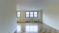 This single-floor space presents a generously sized open living area that immediately draws attention with its distinctive checkered parquet flooring, finished to a polished sheen that reflects natural light throughout the room. The flooring pattern alternates in a classic square design, adding a timeless architectural character to the interior.

Three large windows line the far wall, fitted with dark-framed casings that provide a strong visual contrast against the neutral, cream-colored walls. These windows allow a considerable amount of natural light to fill the space, contributing to an open and airy atmosphere. Below each window, traditional cast-iron radiator units are positioned along the perimeter, indicating a classic heating system consistent with older, well-built construction.

The walls are smooth and painted in a clean off-white tone, complemented by white baseboards and a flat white ceiling. The room's layout is entirely open, offering flexibility for a variety of furniture arrangements. The overall ambiance is bright, understated, and functional, with architectural details that suggest a well-maintained, classically constructed building.