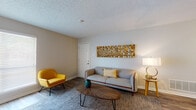 This single-floor residence offers a thoughtfully designed layout with a cohesive mid-century modern aesthetic carried throughout. The living room serves as a welcoming focal point, featuring wood-look flooring, a textured area rug, and warm neutral walls accented by a distinctive mosaic wall art piece. A comfortable sofa, bold yellow accent chair, and an organic-shaped wooden coffee table complete the space with stylish functionality. A window allows natural light to brighten the room, while a media console with barn-door styling provides practical storage. The adjacent dining area is bright and open, with pendant lighting overhead and framed artwork on the walls, flowing seamlessly into the kitchen. The kitchen is outfitted with crisp white shaker-style cabinetry, granite-look countertops, and stainless steel appliances including a range, microwave, and refrigerator, all set atop durable wood-look flooring. The bedroom is a serene retreat with a ceiling fan, a large window with natural light, and direct access to the en-suite bathroom. The bathroom features white cabinetry with brushed gold hardware, granite-look countertops, and a shower/tub combination, maintaining the clean and modern design found throughout the entire residence.