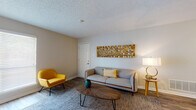 This single-floor residence offers a thoughtfully designed layout with a cohesive mid-century modern aesthetic carried throughout. The living room serves as a welcoming focal point, featuring wood-look flooring, a textured area rug, and warm neutral walls accented by a distinctive mosaic wall art piece. A comfortable sofa, bold yellow accent chair, and an organic-shaped wooden coffee table complete the space with stylish functionality. A window allows natural light to brighten the room, while a media console with barn-door styling provides practical storage. The adjacent dining area is bright and open, with pendant lighting overhead and framed artwork on the walls, flowing seamlessly into the kitchen. The kitchen is outfitted with crisp white shaker-style cabinetry, granite-look countertops, and stainless steel appliances including a range, microwave, and refrigerator, all set atop durable wood-look flooring. The bedroom is a serene retreat with a ceiling fan, a large window with natural light, and direct access to the en-suite bathroom. The bathroom features white cabinetry with brushed gold hardware, granite-look countertops, and a shower/tub combination, maintaining the clean and modern design found throughout the entire residence.