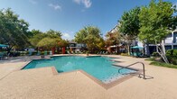 Located in Baton Rouge, Louisiana, this outdoor pool area is a well-appointed amenity space designed for relaxation and recreation. The centerpiece is a generously sized, geometric swimming pool with clear blue water, bordered by a wide, textured concrete sun deck that provides ample space for lounging. The pool's angular shape adds visual interest, while stainless steel entry rails ensure convenient access.

Surrounding the pool, mature trees offer natural shade and a sense of privacy, complementing the verdant landscaping throughout the area. Rows of lounge chairs and teal-accented seating are arranged along the deck, with teal umbrellas providing additional shade near gathering areas. The overall atmosphere is lively yet comfortable, well-suited to the Louisiana climate.

The pool area is framed by brick-clad structures featuring white-trimmed windows with plantation shutters, reflecting a classic Southern architectural style. A covered area adjacent to the pool houses a vending machine station for added convenience. The entrance to the adjacent building features glass double doors beneath a covered walkway, maintaining an inviting, accessible feel. Together, these elements create a cohesive, resort-inspired outdoor environment.