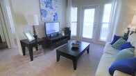 This single-floor living space offers a welcoming and functional layout centered around a well-lit living area. The room is finished with neutral-toned carpet that adds warmth underfoot, while the soft beige walls create a clean, versatile backdrop. Large windows with horizontal blinds flank a glass-panel exterior door, allowing generous amounts of natural light to fill the space and providing a sense of openness. The living area is arranged to maximize comfort, featuring a low-profile dark wood media console that houses a flat-screen television, complemented by matching dark wood accent furniture including a rectangular coffee table and side table. Decorative touches, such as a bold blue floral canvas above the media console and a small potted plant nearby, add personality to the room. The space feels cohesive and thoughtfully organized, with the furniture arrangement encouraging a comfortable viewing and conversational setting. The overall ambiance is bright, calm, and practical, making it a functional everyday living environment.
